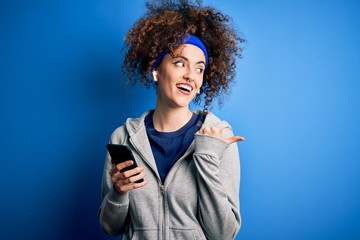 Beautiful sportswoman with curly hair and piercing wearing sportswear using smartphone pointing and showing with thumb up to the side with happy face smiling