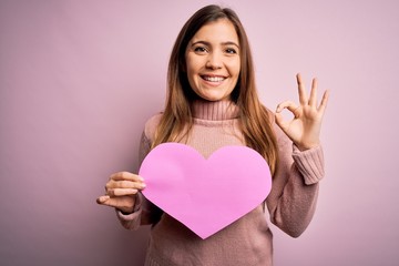 Sticker - Young blonde woman holding romantic big paper heart shape over pink isolated background doing ok sign with fingers, excellent symbol