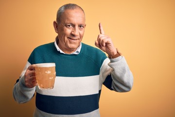 Sticker - Senior handsome man drinking jar of beer standing over isolated yellow background smiling amazed and surprised and pointing up with fingers and raised arms.