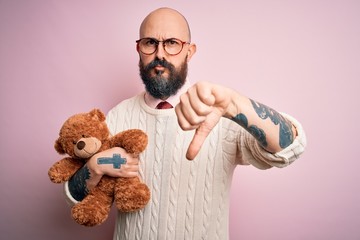 Sticker - Handsome bald man with beard and tattoo holding teddy bear over isolated pink background with angry face, negative sign showing dislike with thumbs down, rejection concept