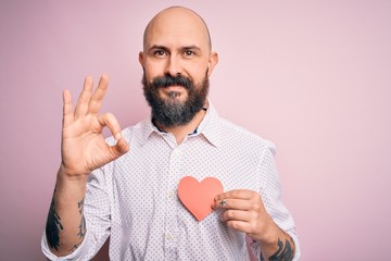 Handsome romantic bald man with beard holding red heart paper over pink background doing ok sign with fingers, excellent symbol