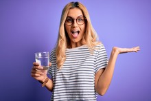 Young Blonde Healthy Woman Wearing Glasses Drinking Glass Of Water Over Purple Background Very Happy And Excited, Winner Expression Celebrating Victory Screaming With Big Smile And Raised Hands