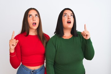 Young beautiful women wearing casual clothes standing over isolated white background amazed and surprised looking up and pointing with fingers and raised arms.