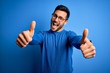 © Krakenimages.com - Young handsome man with beard wearing casual sweater and glasses over blue background approving doing positive gesture with hand, thumbs up smiling and happy for success. Winner gesture.