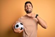 Handsome player man with beard playing soccer holding footballl ball over yellow background cutting throat with hand as knife, threaten aggression with furious violence