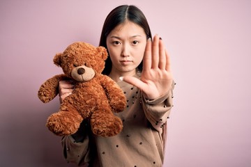 Canvas Print - Young asian woman hugging teddy bear stuffed animal over pink isolated background with open hand doing stop sign with serious and confident expression, defense gesture