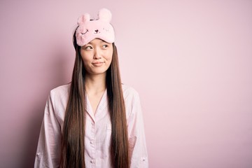 Sticker - Young asian woman wearing pajama and sleep mask over pink isolated background smiling looking to the side and staring away thinking.