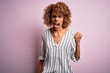 Beautiful african american woman with curly hair wearing striped t-shirt over pink background angry and mad raising fist frustrated and furious while shouting with anger. Rage and aggressive concept.