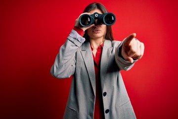 Young beautiful woman with blue eyes using binoculars over isolated red background pointing with finger to the camera and to you, hand sign, positive and confident gesture from the front