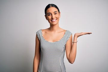 Young beautiful brunette woman wearing casual striped dress over isolated white background smiling cheerful presenting and pointing with palm of hand looking at the camera.