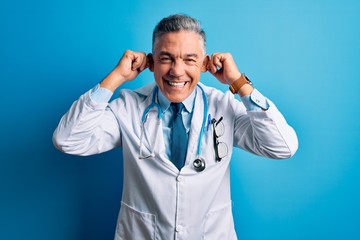 Middle age handsome grey-haired doctor man wearing coat and blue stethoscope Smiling pulling ears with fingers, funny gesture. Audition problem