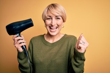 Sticker - Young blonde woman with short hair drying her hair using hairdryer over yellow background screaming proud and celebrating victory and success very excited, cheering emotion