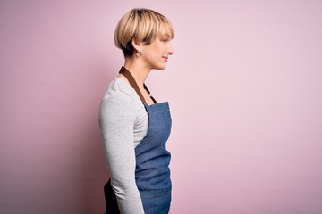 Poster - Young blonde cleaner woman with short hair wearing apron and gloves over pink background looking to side, relax profile pose with natural face with confident smile.