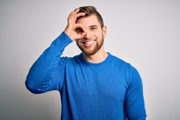 Young handsome blond man with beard and blue eyes wearing casual sweater doing ok gesture with hand smiling, eye looking through fingers with happy face.