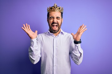 Sticker - Young handsome man with beard wearing golden crown of king over purple background celebrating mad and crazy for success with arms raised and closed eyes screaming excited. Winner concept