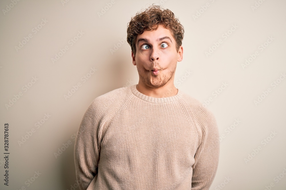 Young blond handsome man with curly hair wearing casual sweater over white background making fish face with lips, crazy and comical gesture. Funny expression.