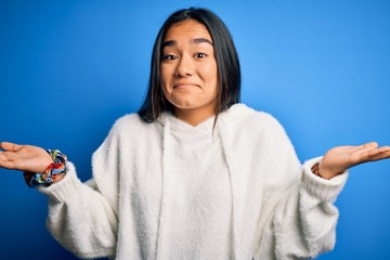 Sticker - Young beautiful asian sportswoman wearing sweatshirt standing over isolated blue background clueless and confused expression with arms and hands raised. Doubt concept.