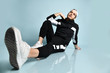 © FAB.1 - Hipster person in cap, black tracksuit, bracelet and sneakers. He is sitting on floor, posing against blue background. Close up