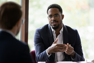 Wall Mural - Confident concentrated African American male employee talk with colleague explain thought or idea, focused biracial businessman speak with coworker or partner, brainstorm at office boardroom meeting