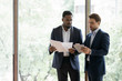 © fizkes - Concentrated diverse businessmen stand near window brainstorm discuss company paperwork together, serious multiracial business partners cooperate consider startup project using tablet gadget