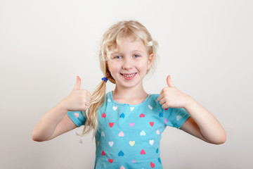 Wall Mural - Portrait of beautiful little funny joyful blonde Caucasian girl on a light background. Happy smiling preschool child kid showing like thumb fingers. Positive emotion face expression.