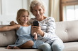 © fizkes - Smiling senior grandmother relax on couch with cute small granddaughter watch funny video on smartphone together, happy granny and little grandchild rest at home enjoy family weekend using cell