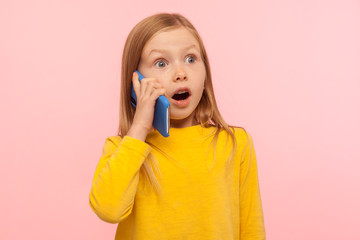 Amazed little ginger girl with freckles calling parents on phone and hearing shocking news, good cellular, roaming, comfortable to use children mobile device. studio shot isolated on pink background