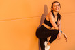 © khosrork - Attractive sexy girl with fit body in tight sportswear, black pants and top, leaning against orange wall and smiling to camera, living healthy sport life, full of energy. isolated on advertising area