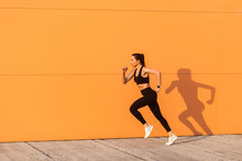 Motivated Confident Fit Woman Athlete In Tight Sportswear, Black Pants And Top, Starting To Run, Jogging Outdoor Against Orange Wall, Advertising Area. Health Care And Weight Loss, Sport Activity