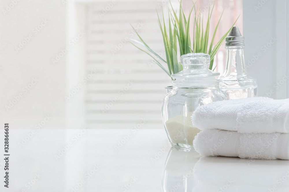 White folded towels and glass bottle on white table with copy space on blurred bathroom background. - obrazy, fototapety, plakaty 