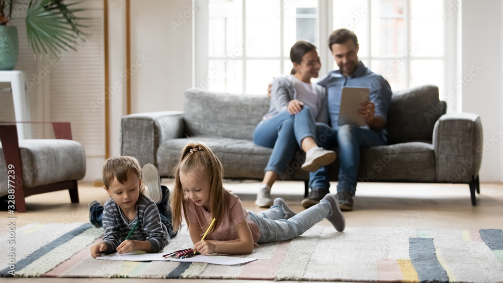 Full length small playful boy lying on floor carpet with happy elder sister, drawing pictures in paper album, while smiling parents resting on sofa, using digital tablet, watching funny videos. - obrazy, fototapety, plakaty 