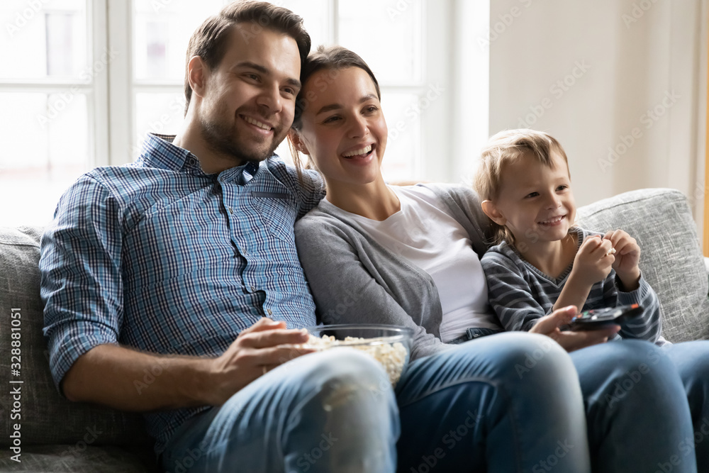 Smiling young man cuddling attractive wife and happy little preschool kid boy, relaxing on comfy sofa, watching show on tv. Joyful family couple enjoying leisure weekend time with small child at home. - obrazy, fototapety, plakaty 