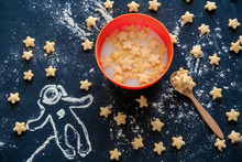 Top View Flat  Bowl Of Milk With Corn Pads In The Shape Of Stars And A Silhouette Of An Astronaut Made Of Flour On A Black Background,play With Food Concept