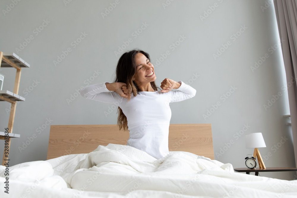 Overjoyed young Caucasian woman sit in cozy white home bedroom wake up stretching feel positive, happy smiling millennial girl awaken do morning exercise in comfortable bed, optimism concept - obrazy, fototapety, plakaty 