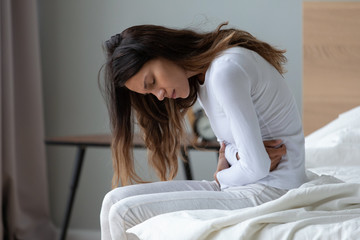 sick young woman sit on bed at home touch belly suffer from stomach ache, stressed ill millennial gi