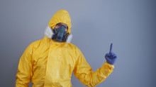 A Man In A Yellow Chemical Protection Suit On A Gray Background Raises His Index Finger Up. Copy Space