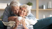 Caring Elderly 60s Husband Caress Hug Middle-aged Wife Bring Tea Cover With Blanket At Home, Happy Old Mature Couple Relax Enjoy Weekend In Living Room Together Show Love And Affection