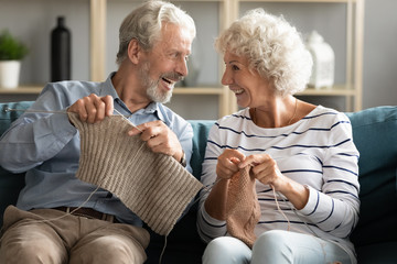 Overjoyed retired old husband and wife sit relax on couch in living room knitting together, happy smiling elderly mature couple spouses engaged in hobby activities enjoy leisure weekend at home