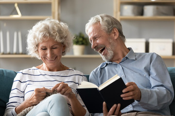 Overjoyed elderly mature couple spouses sit rest on couch in living room engaged in favorite hobby activities together, happy middle-aged retired husband laugh reading funny book, wife do knitting