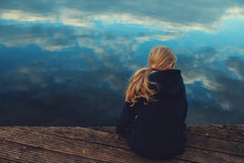 Young Girl With Long Blonde Hair Is Sitting On A Wooden Stage While Looking Over The Rippled Water With Reflecting Cloudy Sky In A Thoughtful Pose - Coming Of Age, Outdoor And Nature Care Concept