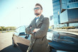 © Fxquadro - Confident businessman standing outdoors in a city near glass building and his car, wearing white shirt, sunglasses, bow tie and dark coat, looking cool and successful