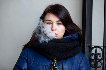 Wall Mural - Vape teenager. Young pretty white caucasian brunette girl with blue eyes and in a blue jacket smoking an electronic cigarette near old metal gate on the street in the spring. Deadly bad habit.