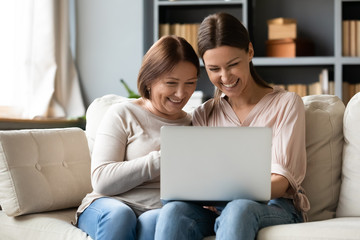 Wall Mural - Happy older mature mother sitting on sofa with excited grownup daughter, looking at laptop screen. Laughing young woman showing funny video photos to elderly mommy, having fun together at home.