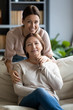 © fizkes - Vertical image portrait happy grownup daughter hugging pleasant older brunette mother resting on couch. Smiling young woman posing for bonding family photo with mom in living room, looking at camera.