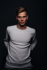 Canvas Print - Informally ( casual ) dressed blonde young man with sharp jawline in his 20's posing in a studio in front of a black background while wearing a white sweater.