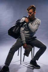 Canvas Print - Informally ( casual ) dressed blonde model wearing a white sweater and holding a black man bag is posing on a white background while sitting on a chair.