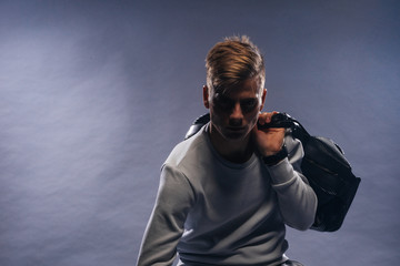 Canvas Print - Informally ( casual ) dressed blonde model wearing a white sweater and holding a black man bag is posing on a white background while sitting on a chair.