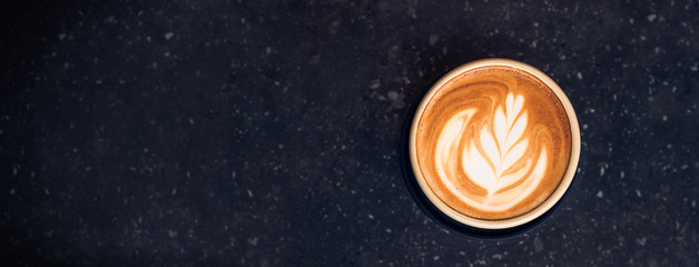 Wall Mural - Top view hot capucino coffee cup with latte art foam on black stone table at cafe.dark scene.banner space mock up for adding text