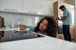 © Svitlana - Professional Service. Young afro american woman with serious face wiping electric stove with textile wipe while working with her male coworker in the modern kitchen