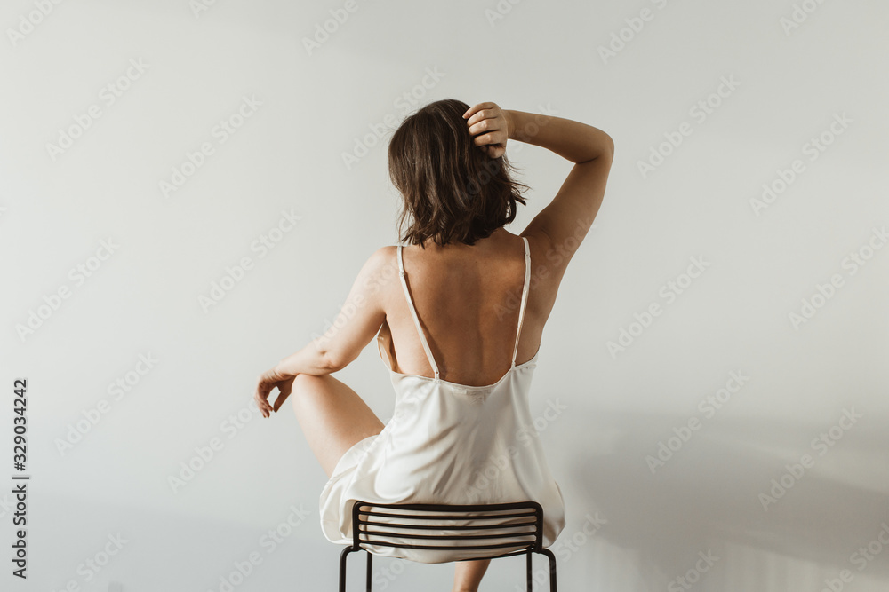 Young pretty woman in silk underwear sitting on black stool on white background. Fashion concept. - obrazy, fototapety, plakaty 